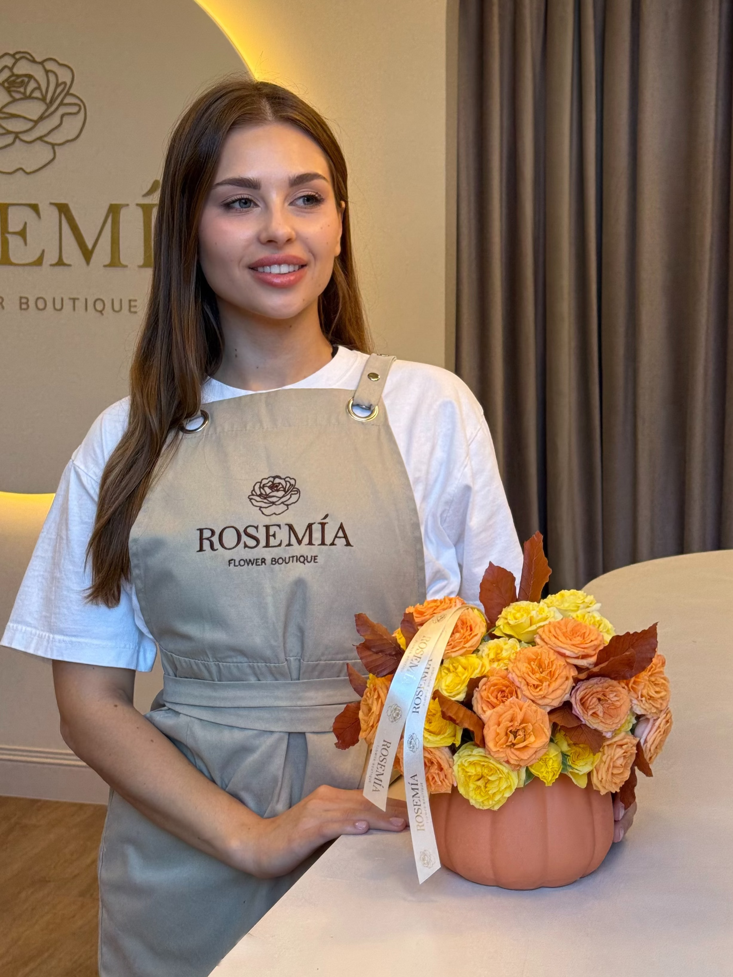 A woman wearing a white t-shirt and a light gray apron with 'ROSEMÍA CLINIC BOUTIQUE' printed on it, holding a small terracotta pot filled with orange, yellow, and peach artificial roses. The background shows a boutique interior with a gray curtain and a logo on the wall.