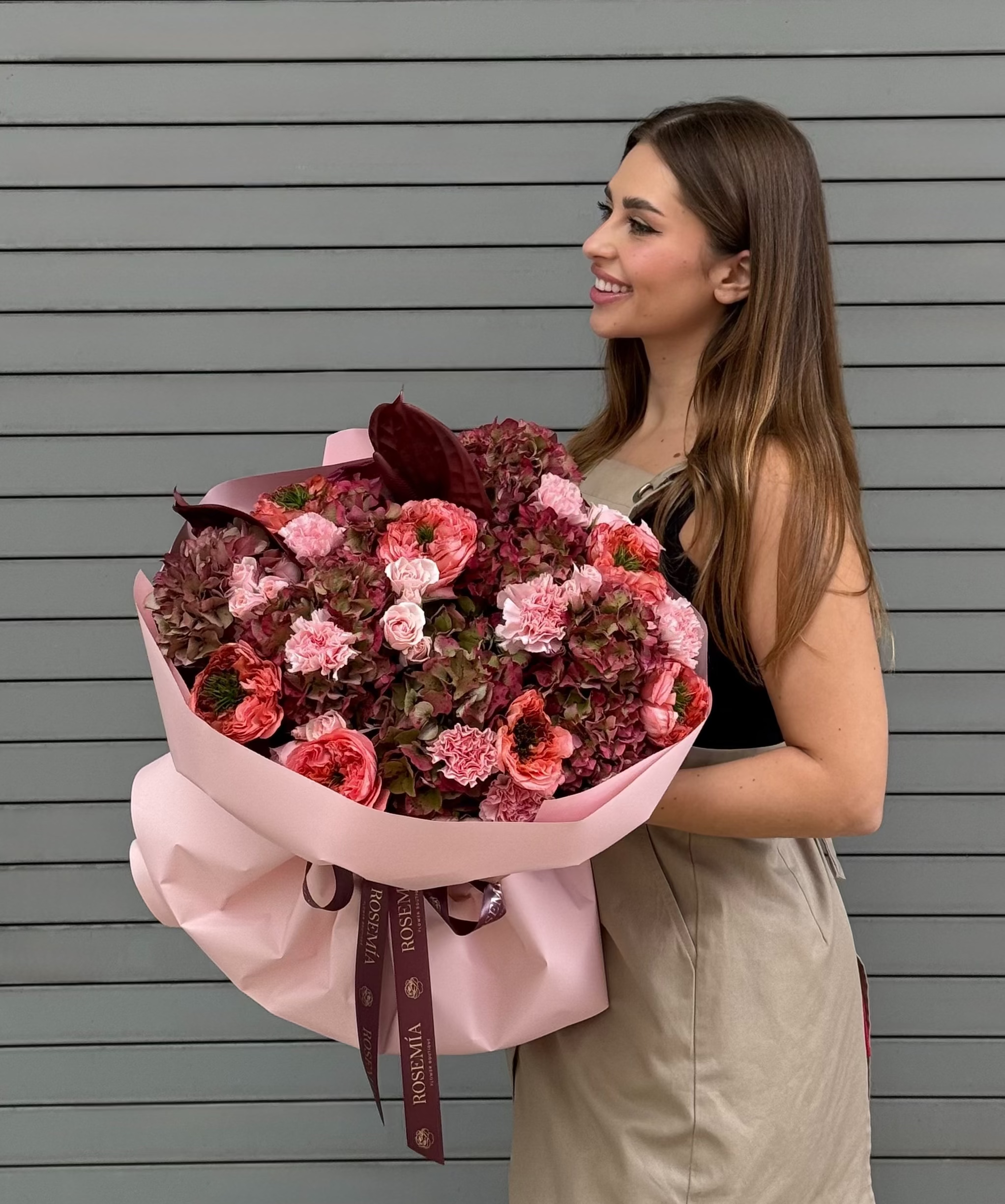 A woman with long brown hair stands in front of a gray horizontal-slat wall, holding a large bouquet of pink, red, and burgundy flowers wrapped in light pink paper with a dark ribbon.