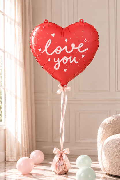 A red heart-shaped foil balloon with 'love you' written in white script, tied to a wrapped gift bag with a pink ribbon, set in a softly lit room with pastel balloons on the floor.