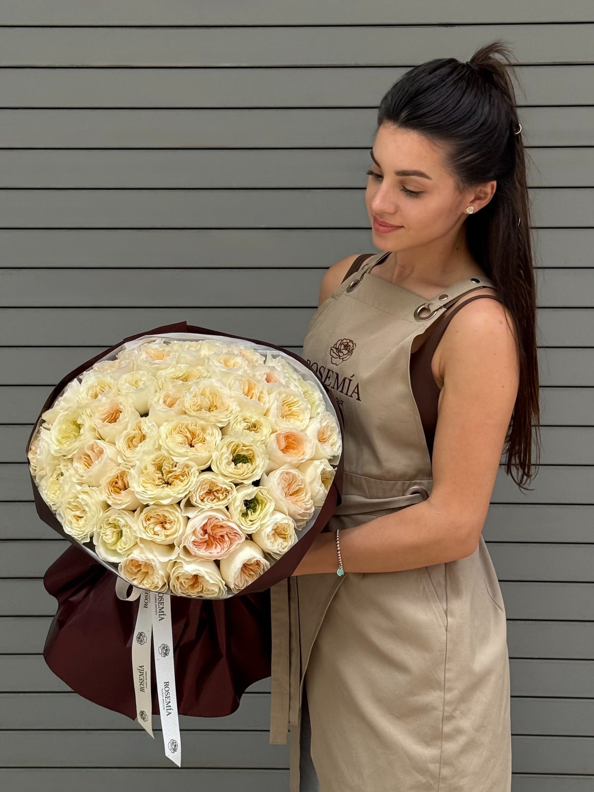 A woman in a beige dress holding a large bouquet of white and pale yellow roses with a dark burgundy wrap, standing against a gray horizontal-slat wall.