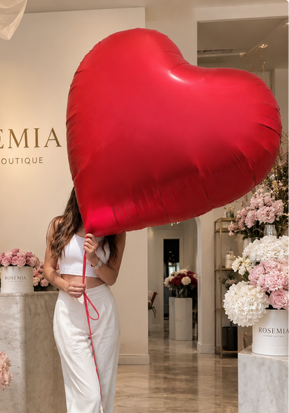 A woman in a white crop top and white pants stands in a flower boutique holding a large red heart-shaped balloon. The boutique interior features marble floors, a reception desk with a laptop, and multiple white flower buckets filled with pink and white roses.