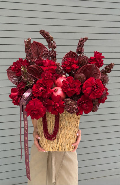 A person holds a large woven basket filled with a dense floral arrangement in rich reds and deep burgundy tones, including flowers and a pomegranate, against a gray slatted wall.