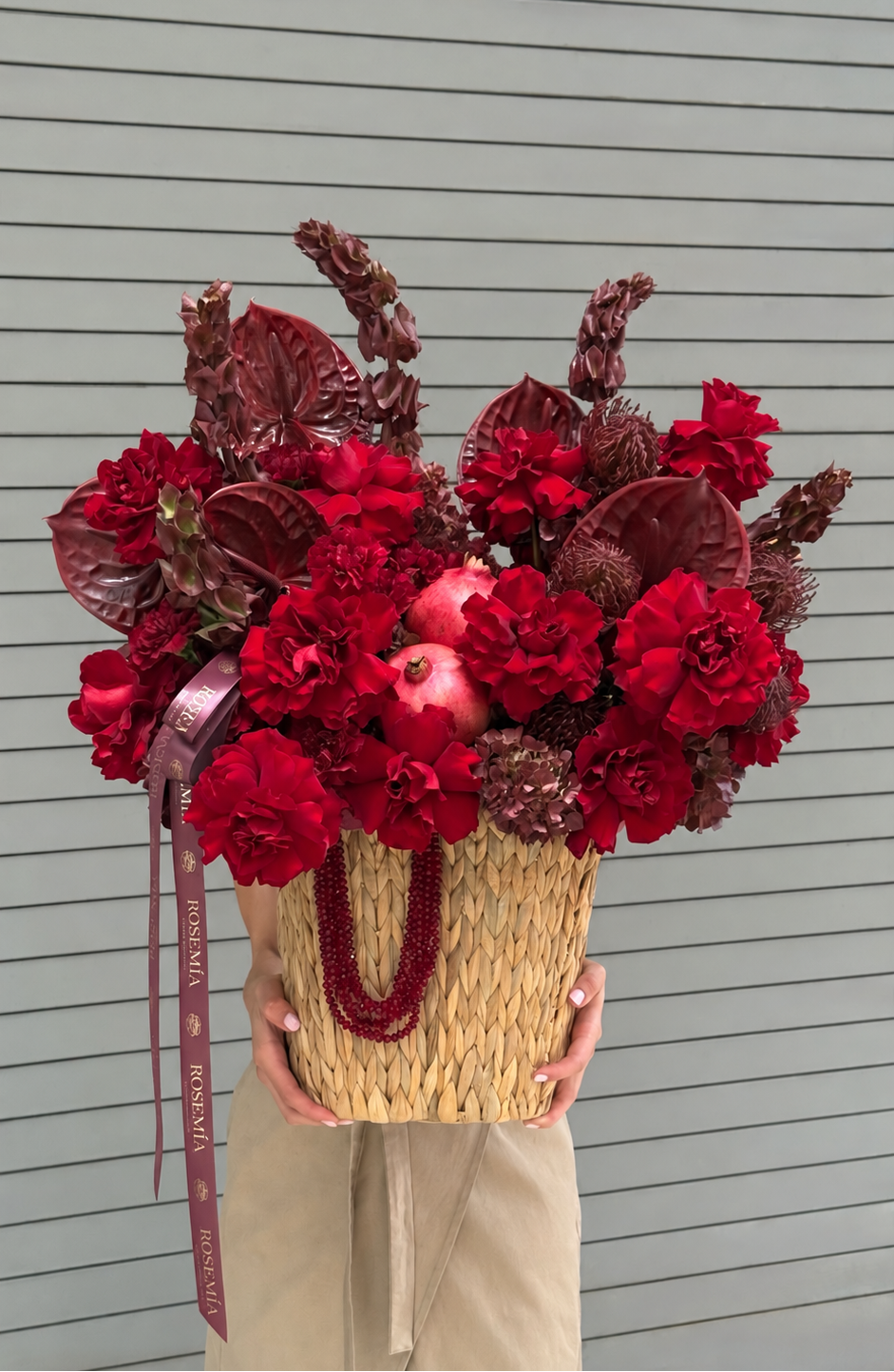 A person holds a large woven basket filled with a dense floral arrangement in rich reds and deep burgundy tones, including flowers and a pomegranate, against a gray slatted wall.