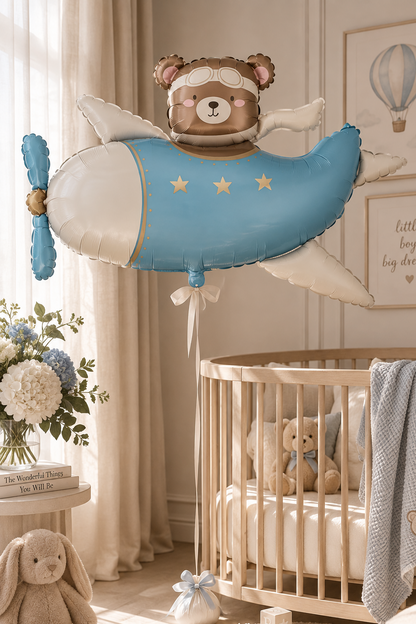 A large decorative balloon shaped like a baby airplane with a teddy bear head and small angel wings, floating in a softly styled nursery with a crib, stuffed bunny, framed wall art, and warm natural light.