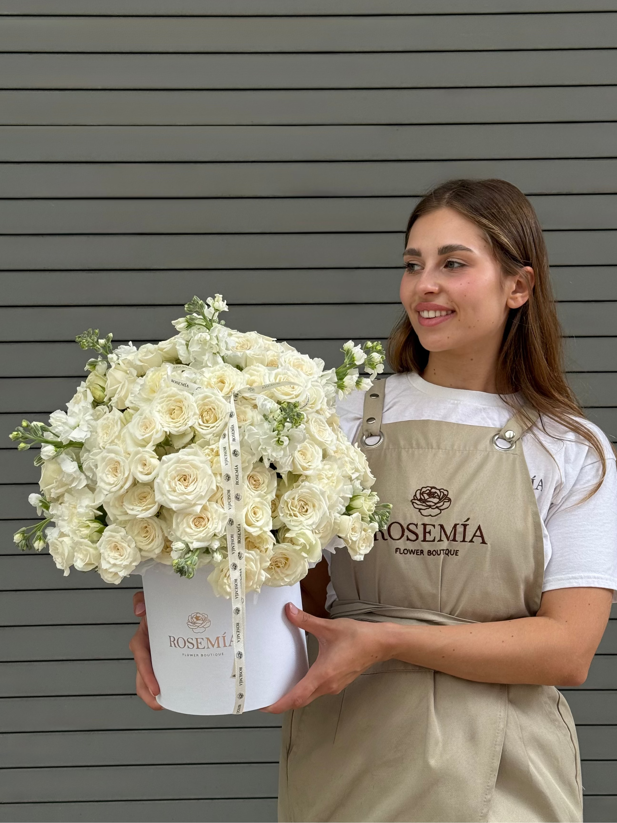 Woman holding a large bouquet of white flowers in front of a gray wall with 'Rosemia' branding.