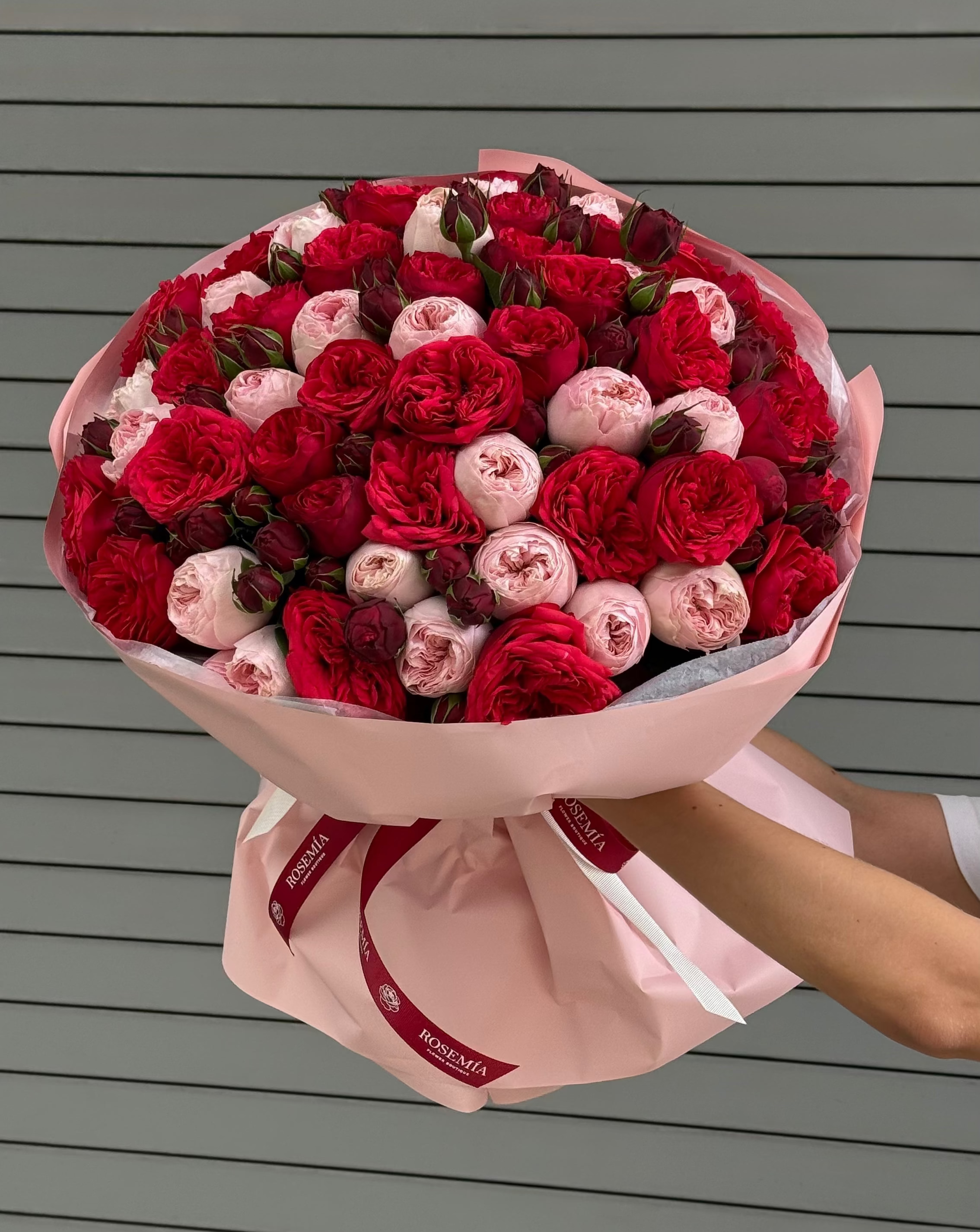 A large bouquet of red and pink roses wrapped in light pink paper with a red ribbon, held by a person against a gray horizontal-slat wall.