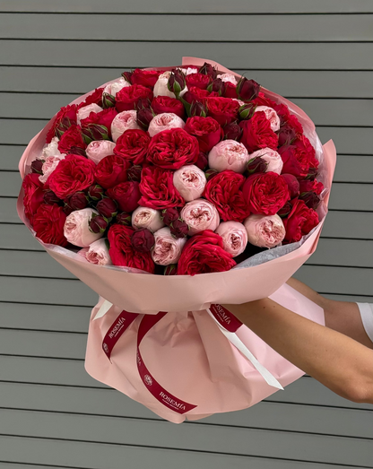 A large bouquet of red and pink roses wrapped in light pink paper with a red ribbon, held by a person against a gray horizontal-slat wall.