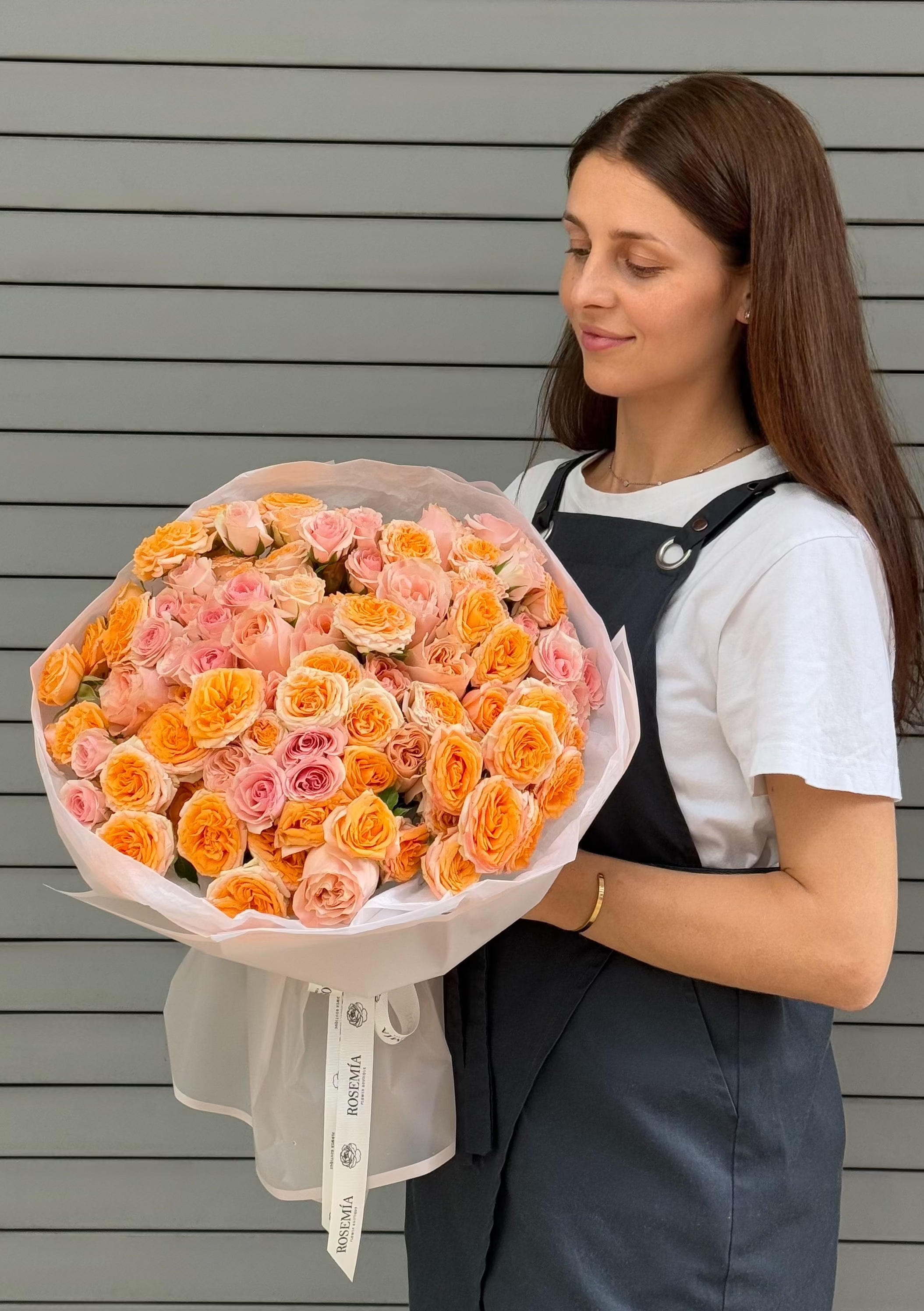 A woman with long brown hair stands in front of a gray slatted wall, holding a large bouquet of orange and pink roses wrapped in white tissue paper.