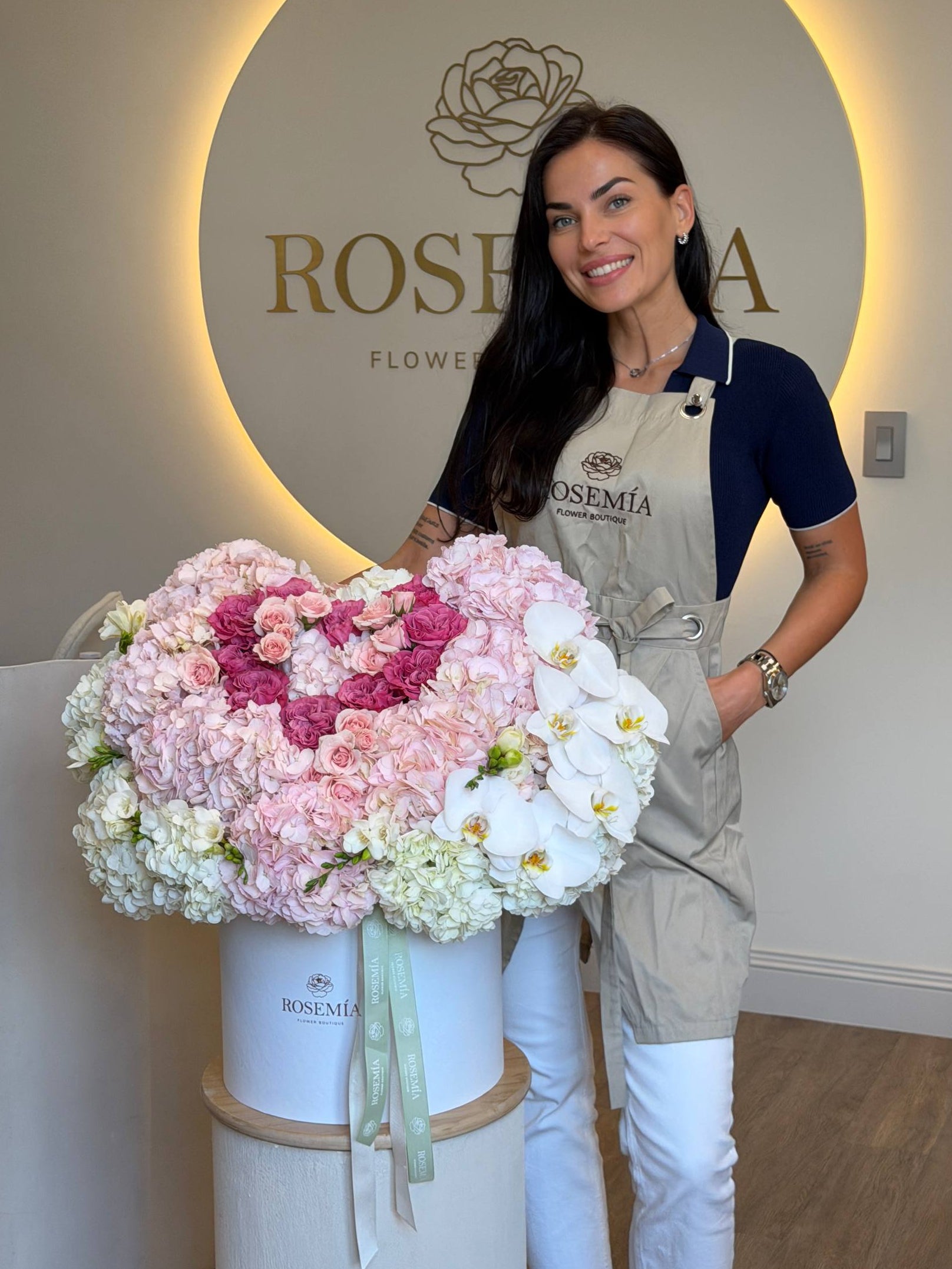 A smiling woman wearing a beige floral apron over a navy shirt and white pants stands behind a large white cylindrical box filled with pink, blush, and white flowers. The background features a warm-lit circular wall sign with the brand name 'ROSEMIA FLOWER'.