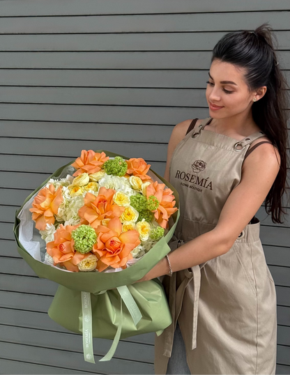 A woman wearing a beige floral arrangement apron holds a large, vibrant bouquet of orange, yellow, and green flowers in front of a gray horizontal-slat wall.