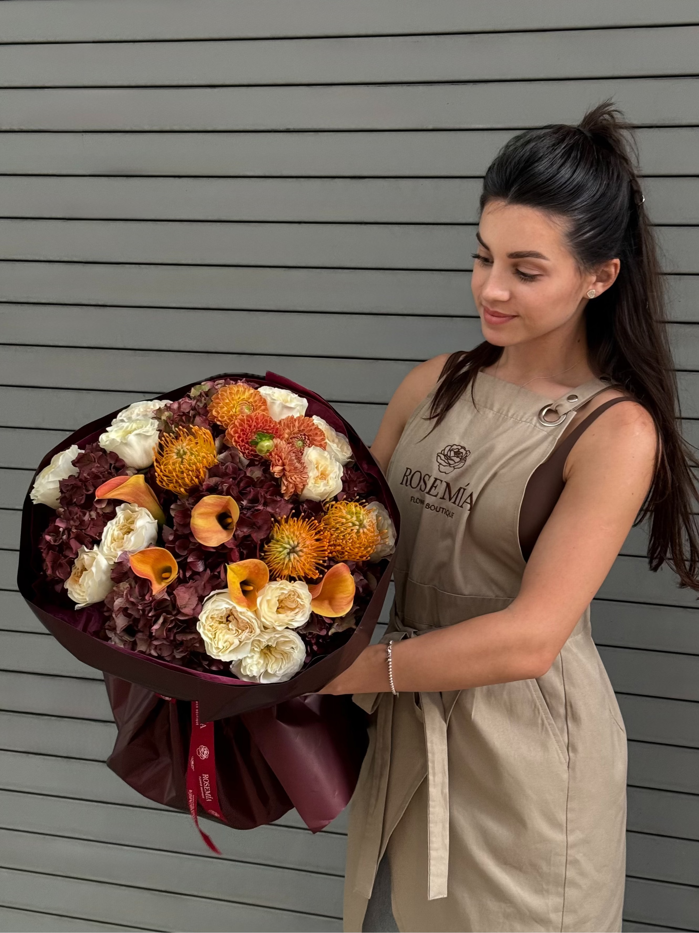 A woman in a beige dress holding a large, colorful flower bouquet with orange, white, and burgundy flowers, standing against a gray horizontal-slat wall.