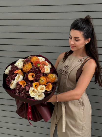 A woman in a beige dress holding a large, colorful flower bouquet with orange, white, and burgundy flowers, standing against a gray horizontal-slat wall.