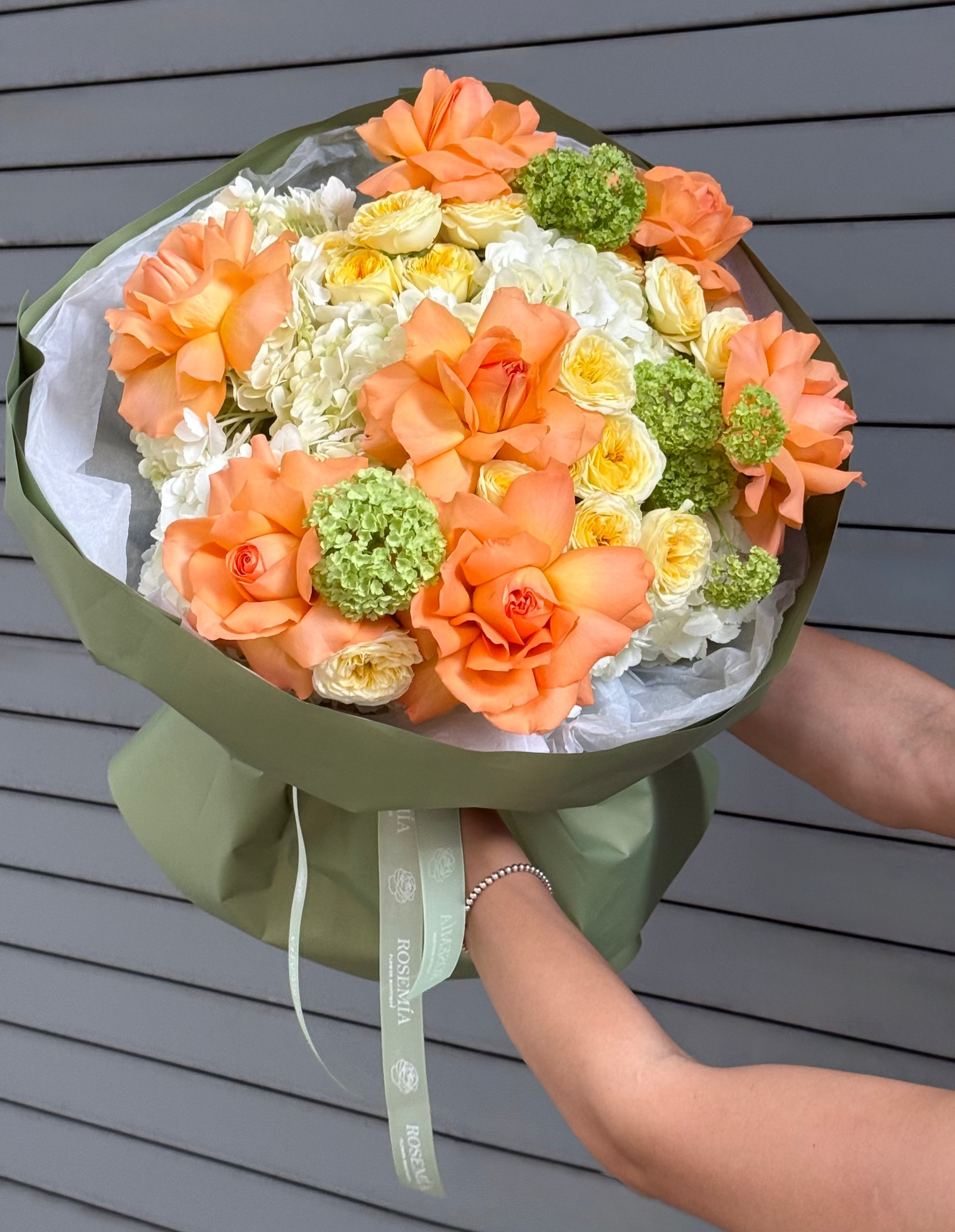 A bouquet of orange carnations, white carnations, and green hydrangea-like flowers wrapped in olive green paper with a white ribbon, held by hands against a gray slatted wall.