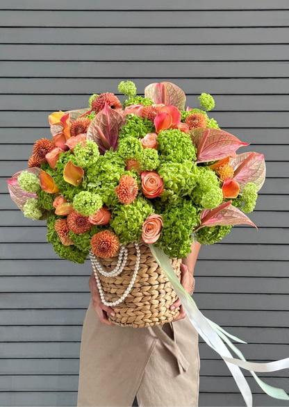 A large, vibrant autumnal flower bouquet held by a person wearing beige trousers, set against a gray horizontal-slat wall. The bouquet features green hydrangeas, orange and pink flowers, and pinkish-red leaves, arranged in a woven straw basket with a pearl chain.