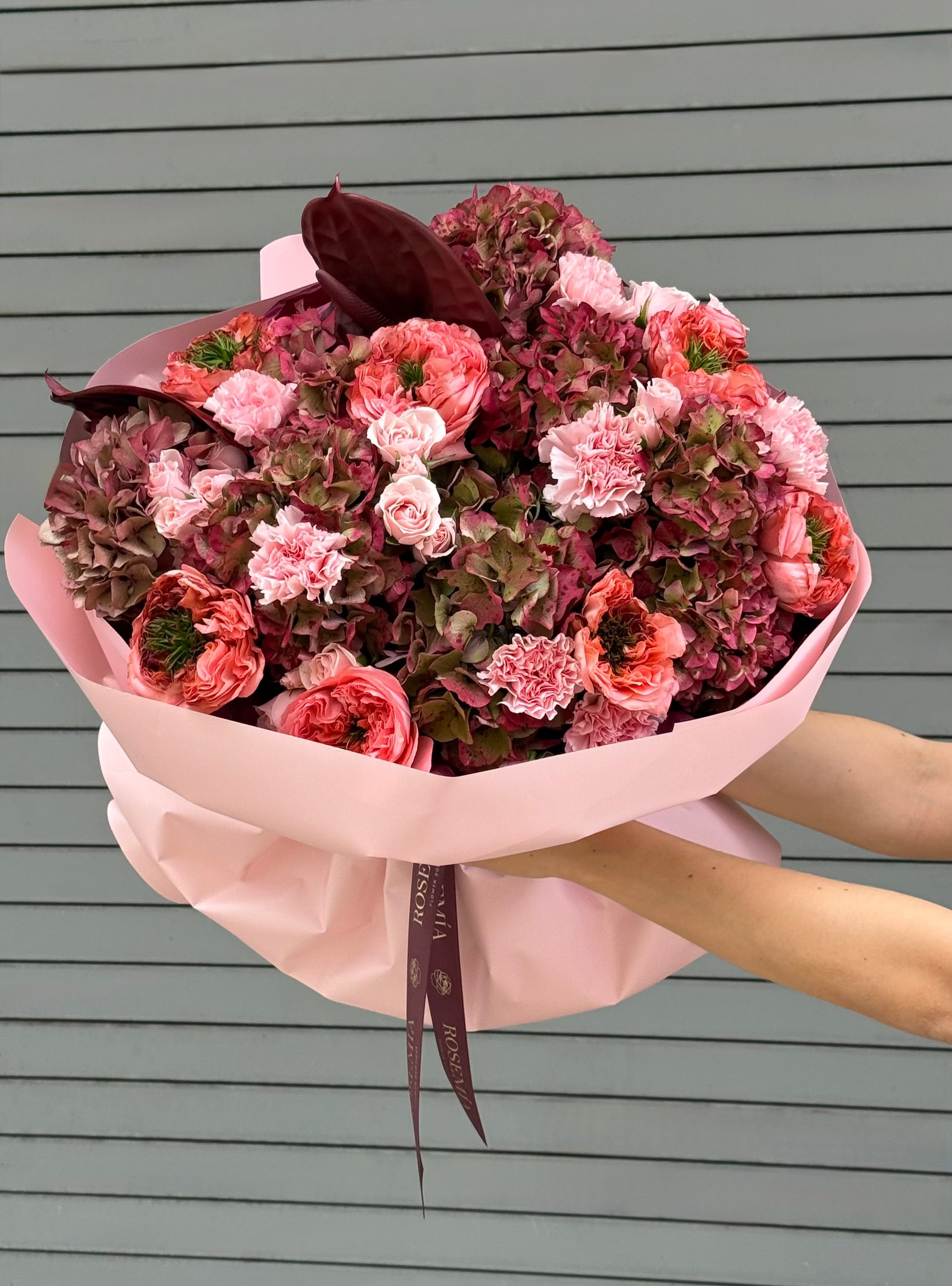 A hand-held bouquet of pink and red flowers wrapped in light pink paper, held by two hands against a gray horizontal-slat background.