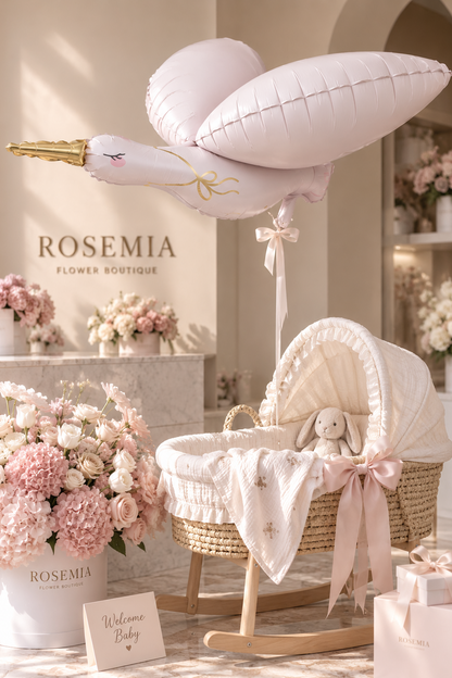 A styled baby nursery scene featuring a woven rattan bassinet with a cream lace canopy and ribbon, a large pale pink bird-shaped balloon, pink floral arrangements, gift boxes, and a sign reading 'Welcome Baby.' The wall displays 'ROSEMIA FLOWER BOUTIQUE,' which appears to be the visible brand.