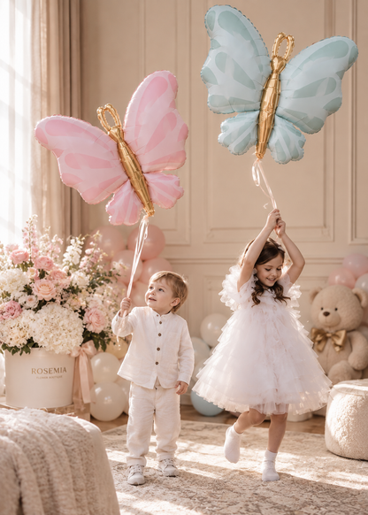 A young child in a white tulle dress stands in a decorated nursery holding a large light blue butterfly balloon, while a toddler in a white outfit holds a large pink butterfly balloon nearby. The room has soft beige walls, a teddy bear, balloons, and floral arrangements.